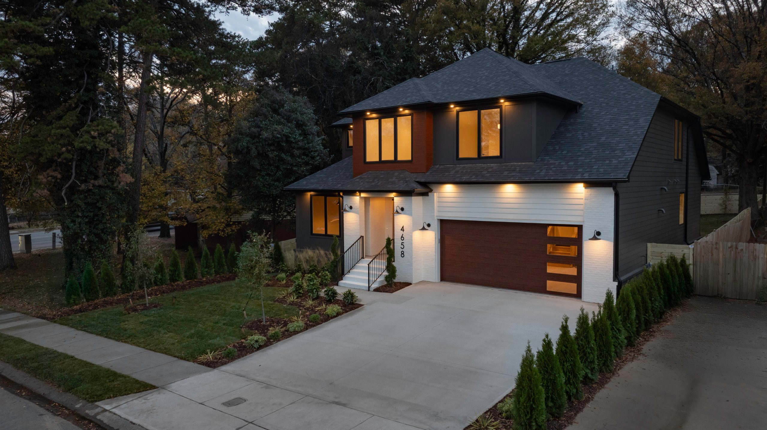 Two-story new construction home with dark charcoal siding, white brick, and wood accents, photographed at dusk with warm interior lighting