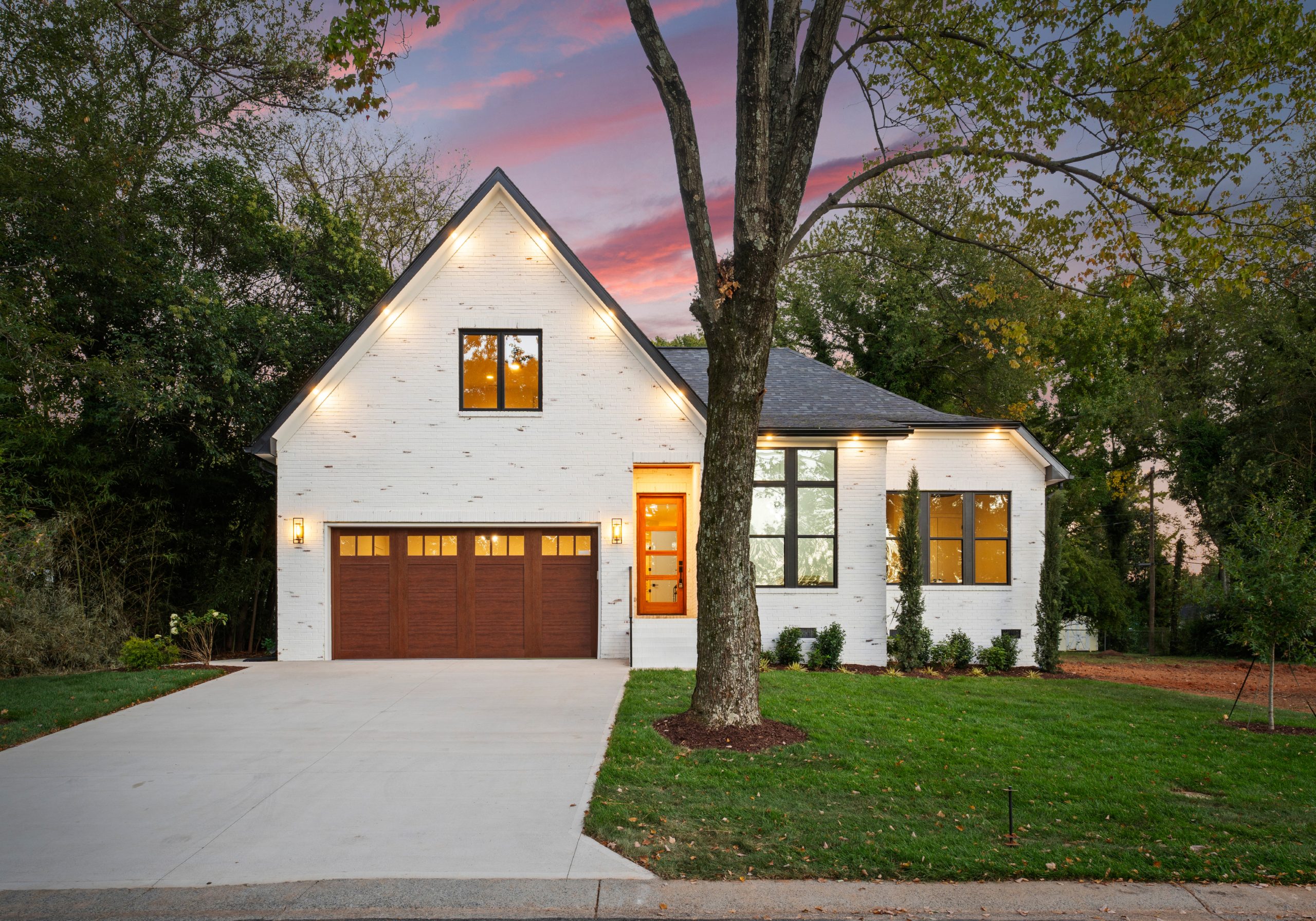 White-painted brick two-story home with steep front gable, orange front door, and string lights along the roofline, photographed at dusk