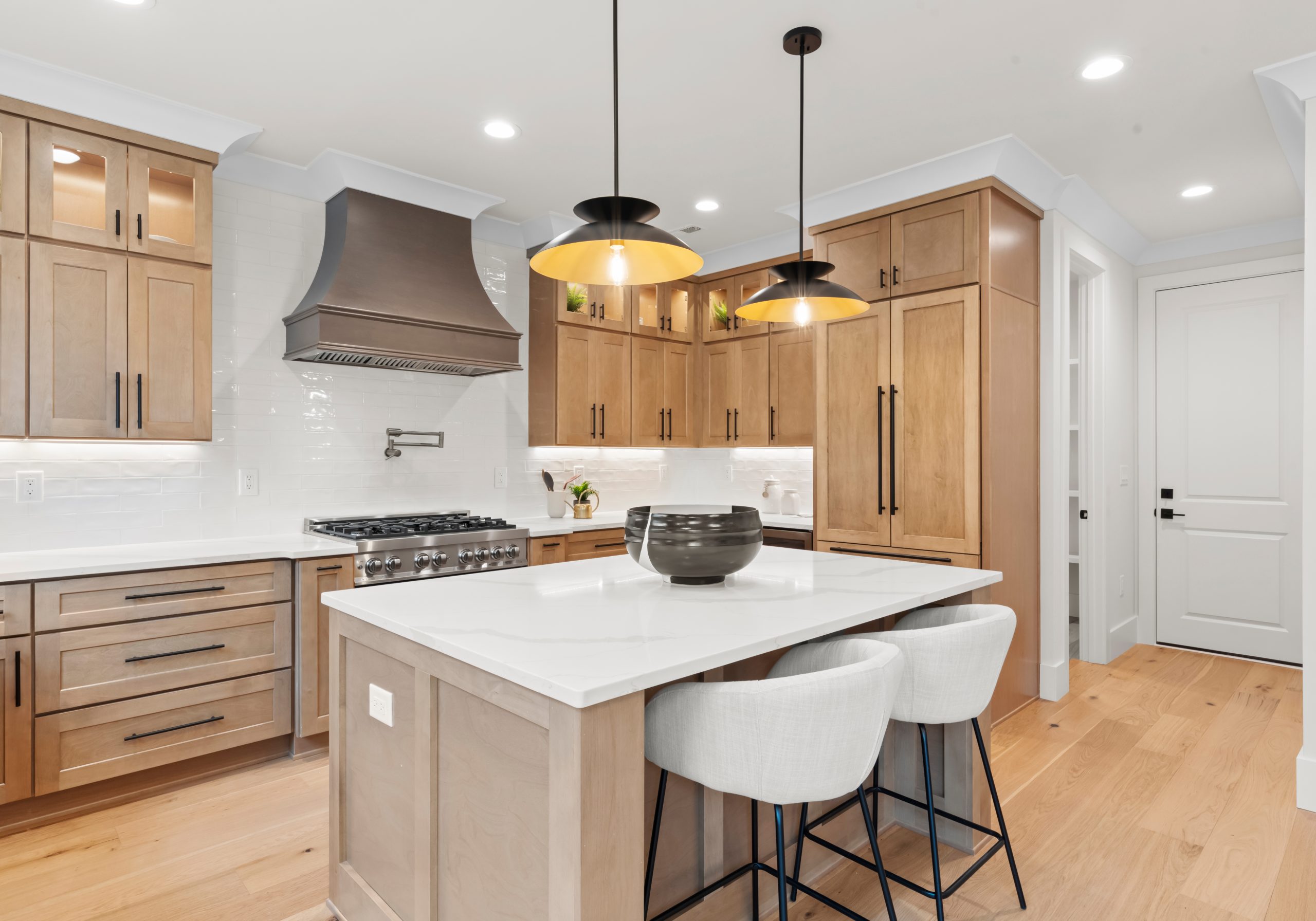 Kitchen with light wood cabinetry, a stainless range hood, quartz island, and black pendant lighting